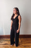A woman modeling a black sleeveless jumpsuit with a V-neck bodice and wide legs, standing on a wooden floor against a plain off-white wall.