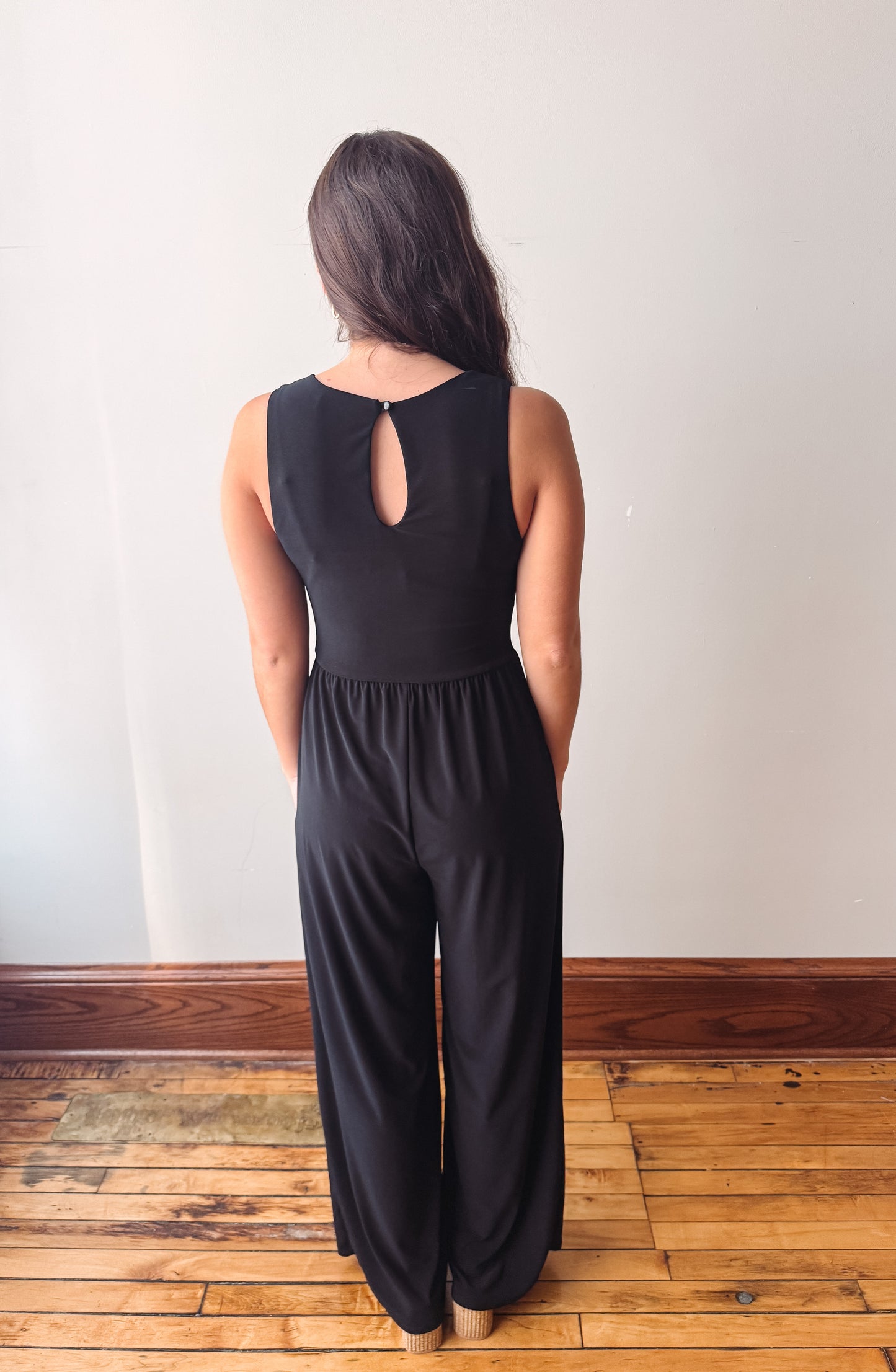 A woman viewed from behind wearing a black sleeveless jumpsuit with a keyhole back cutout, standing on a wooden floor against a plain white wall.