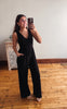 A woman takes a mirror selfie wearing a black sleeveless jumpsuit with a V-neck and wide legs, standing on a wooden floor in front of a plain wall.