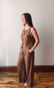 A woman wearing a sleeveless, wide-leg brown jumpsuit with front buttons, standing in profile against a plain off-white wall on a wooden floor.
