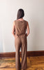 A woman viewed from behind wearing a sleeveless brown jumpsuit with wide-leg pants, standing on a wooden floor against a plain off-white wall.