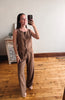 A woman takes a mirror selfie wearing a sleeveless brown vest and matching wide-leg pants set, standing on a wooden floor in a room with a white wall and a small cabinet.