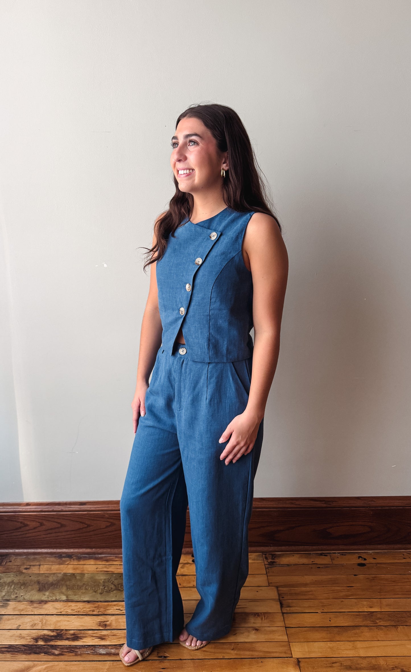 A woman modeling a sleeveless blue wide-leg jumpsuit with decorative buttons down the front, standing on a wooden floor against a plain light gray wall.
