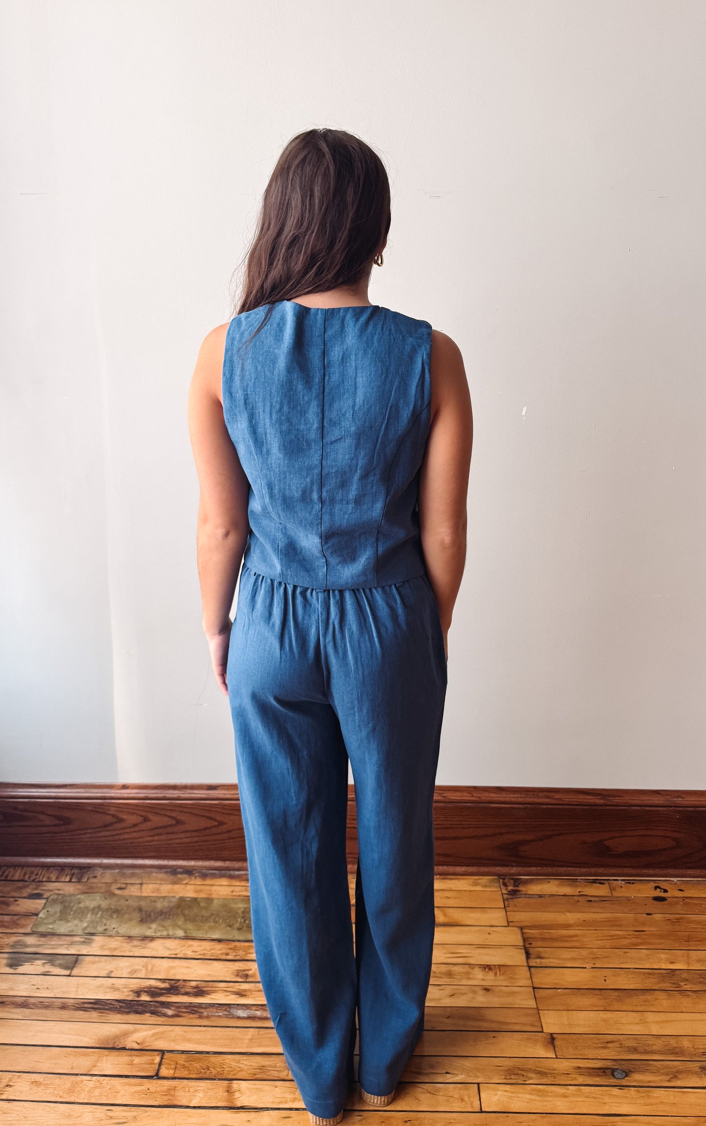 A woman viewed from behind wearing a sleeveless blue jumpsuit with wide legs, standing on a wooden floor against a plain off-white wall.