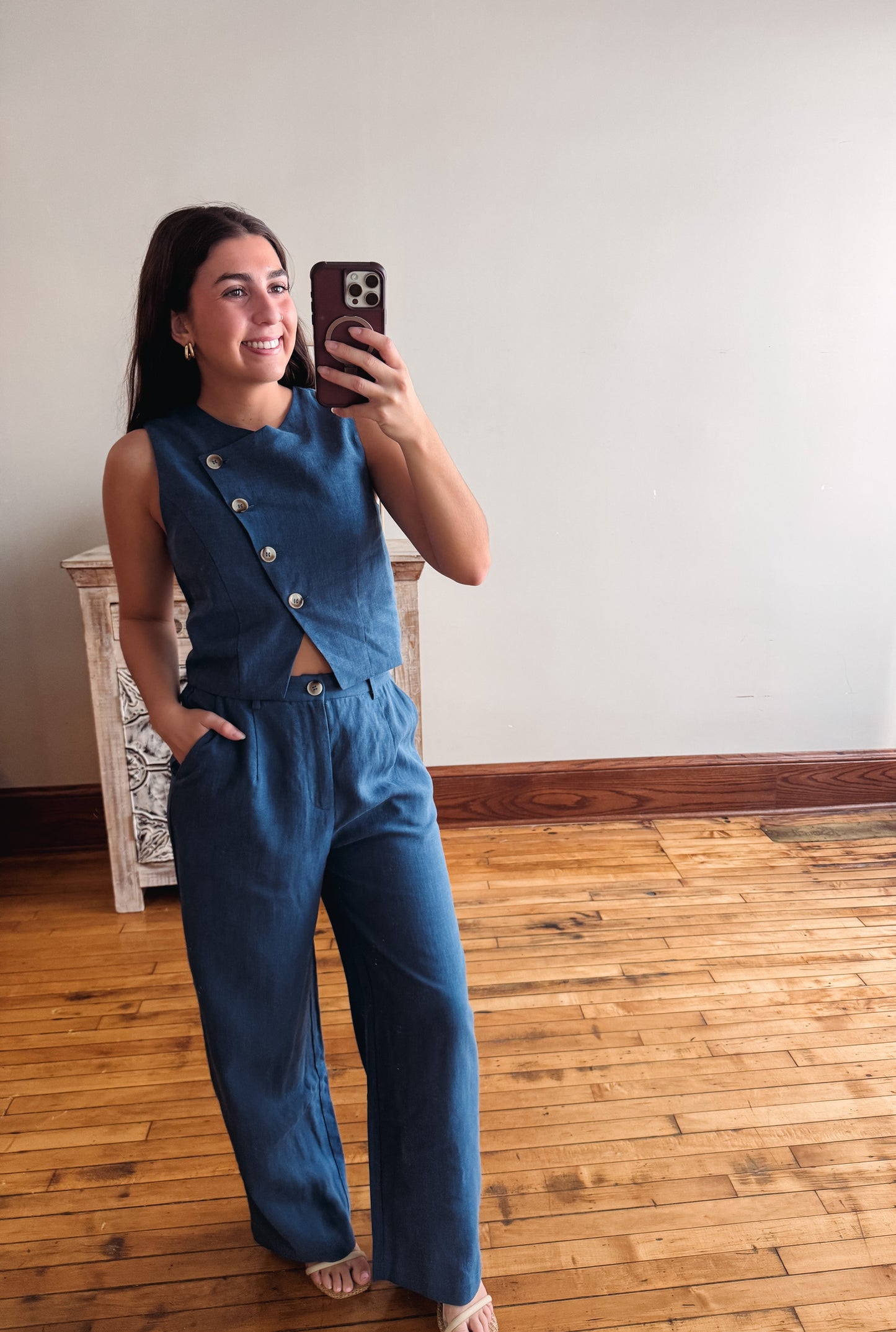 A woman takes a mirror selfie wearing a sleeveless blue denim-like jumpsuit with front buttons and wide legs, standing in a room with a light wall and wood floor.