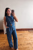 A woman takes a mirror selfie wearing a sleeveless blue denim-like jumpsuit with front buttons and wide legs, standing in a room with a light wall and wood floor.