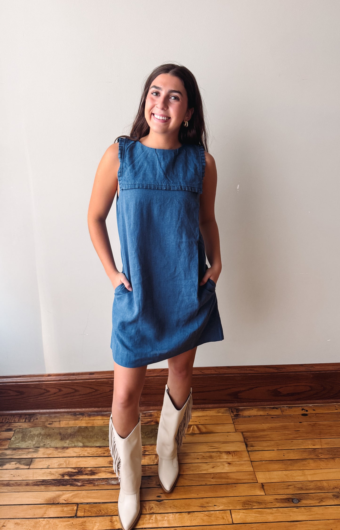 A woman modeling a sleeveless blue denim dress with side pockets, paired with cream-colored cowboy boots, standing on a wooden floor against a plain off-white wall.