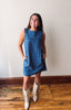 A woman modeling a sleeveless blue denim dress with side pockets, paired with cream-colored cowboy boots, standing on a wooden floor against a plain off-white wall.