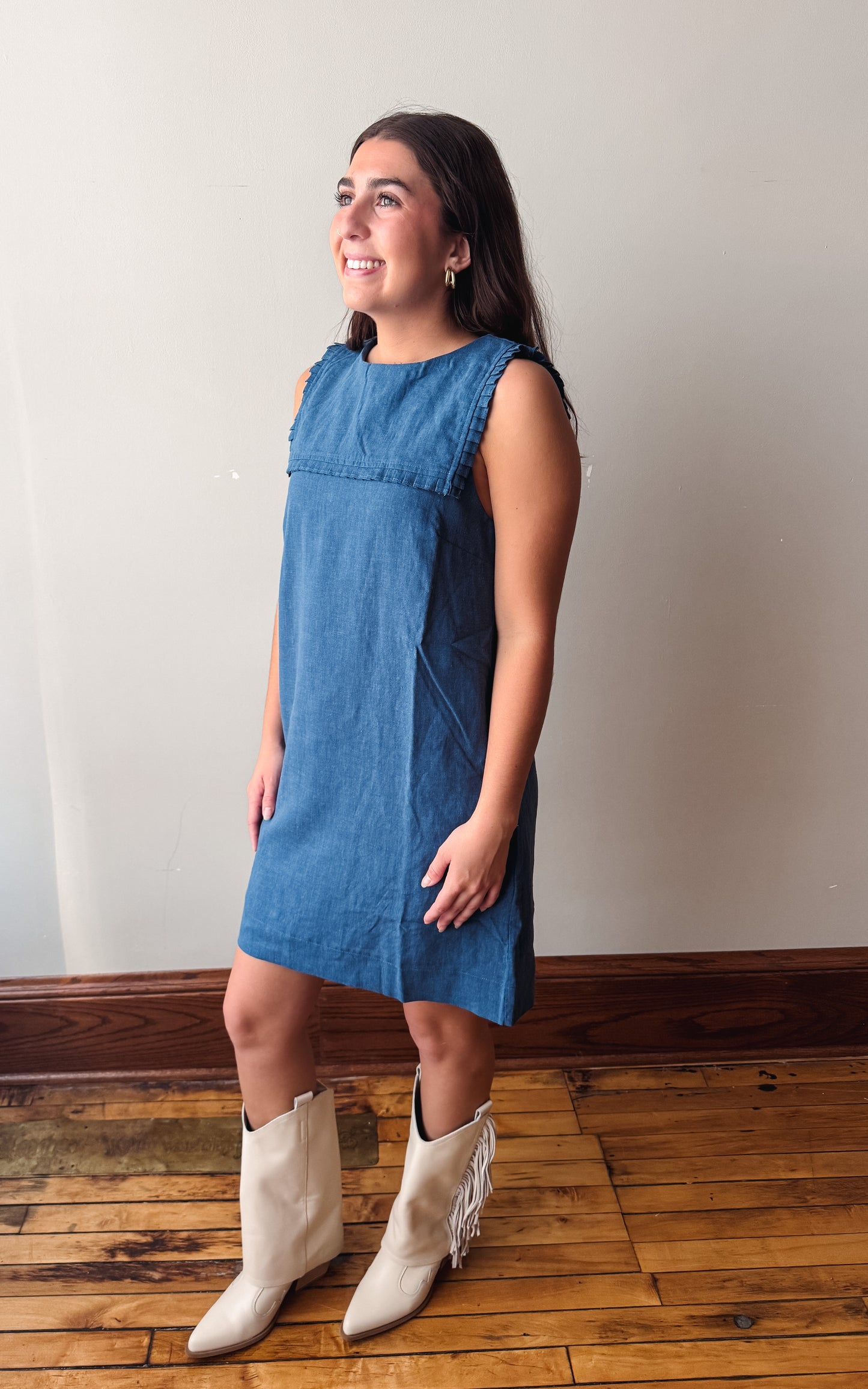A woman modeling a sleeveless blue denim dress with fringe detailing at the neckline, paired with cream-colored fringed ankle boots, standing on a wooden floor against a plain wall.
