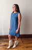 A woman modeling a sleeveless blue denim dress with fringe detailing at the neckline, paired with cream-colored fringed ankle boots, standing on a wooden floor against a plain wall.