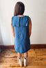 A woman viewed from behind wearing a sleeveless blue denim dress with ruffled shoulder details and cream-colored fringed boots, standing on a wooden floor against a plain white wall.