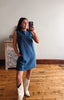 A woman takes a mirror selfie wearing a sleeveless blue denim dress and white cowboy boots in a room with a light gray wall and wooden floor.