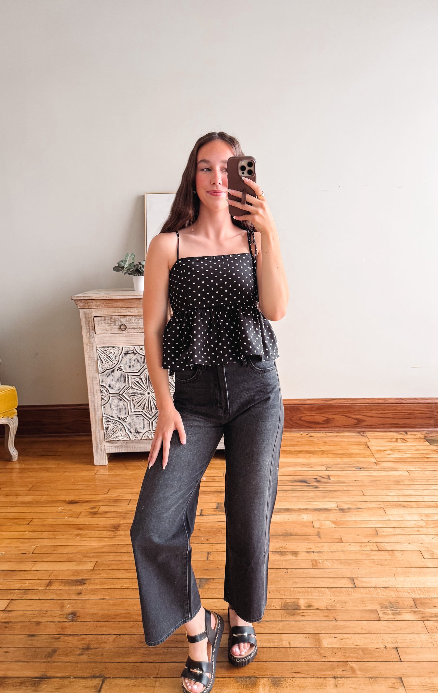 Woman taking a mirror selfie wearing a black polka dot top and gray pants in a room with wooden flooring.