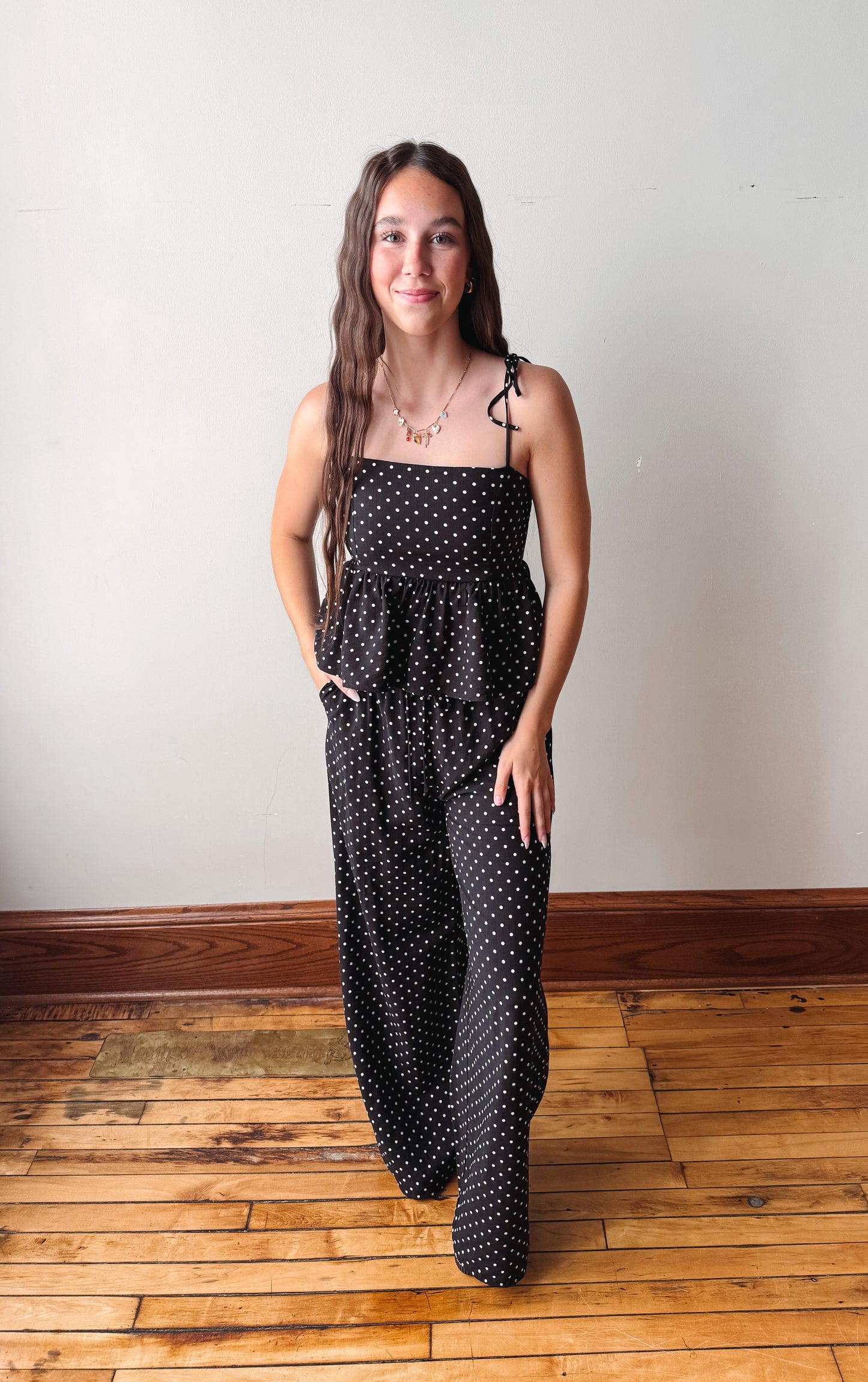 Woman wearing a black polka dot dress standing on a wooden floor with a white wall background