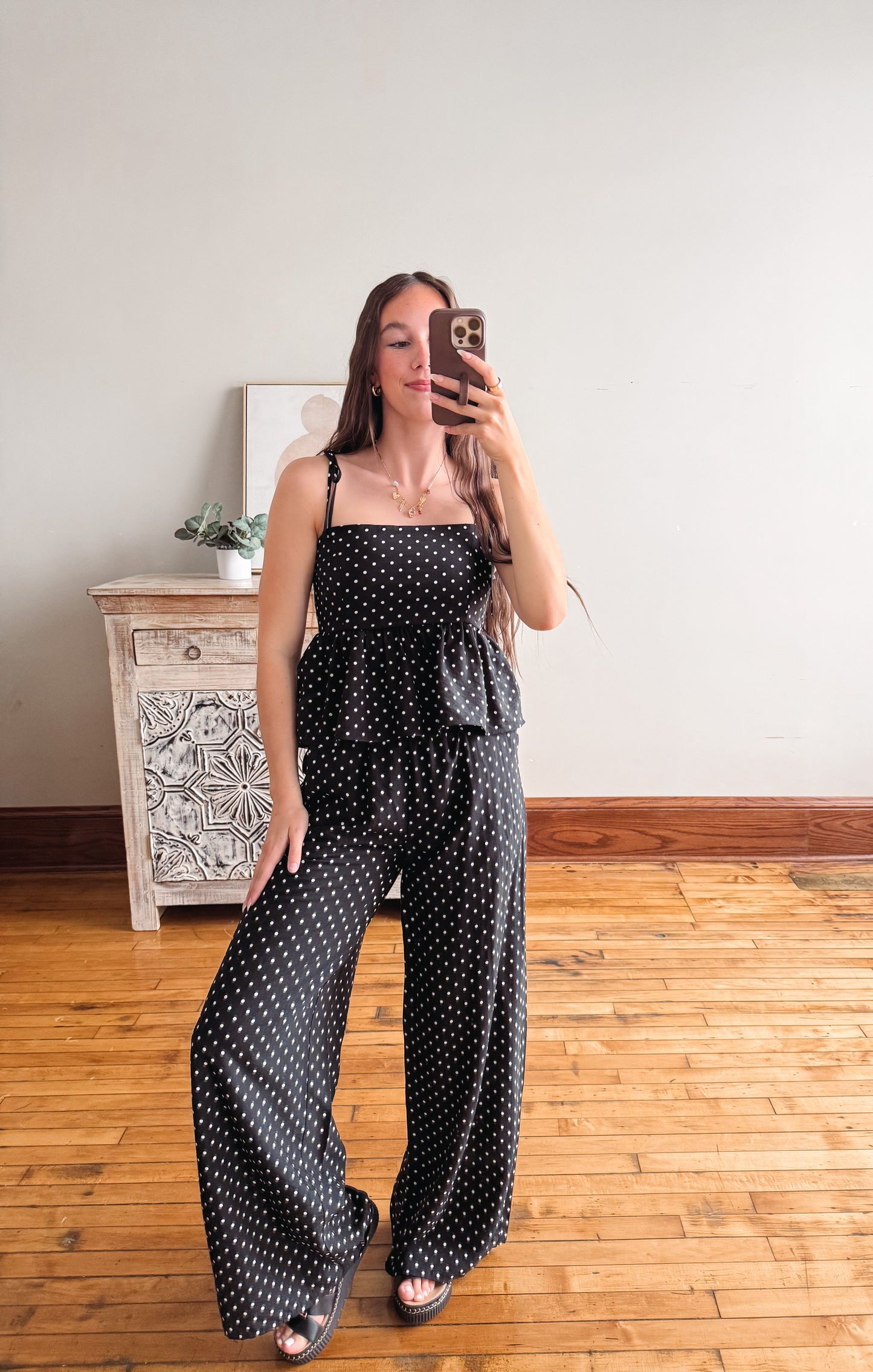 Woman taking a mirror selfie wearing a black polka dot jumpsuit in a room with wooden flooring and a decorative cabinet.
