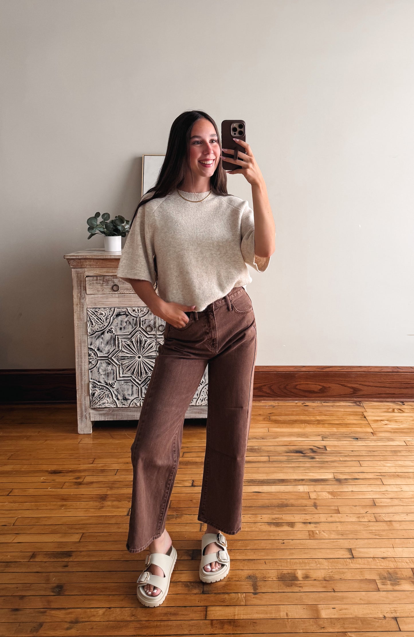 A woman takes a mirror selfie in a room with hardwood floors, wearing a beige short-sleeve knit top and brown wide-leg cropped pants with white slide sandals.