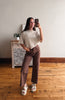 A woman takes a mirror selfie in a room with hardwood floors, wearing a beige short-sleeve knit top and brown wide-leg cropped pants with white slide sandals.