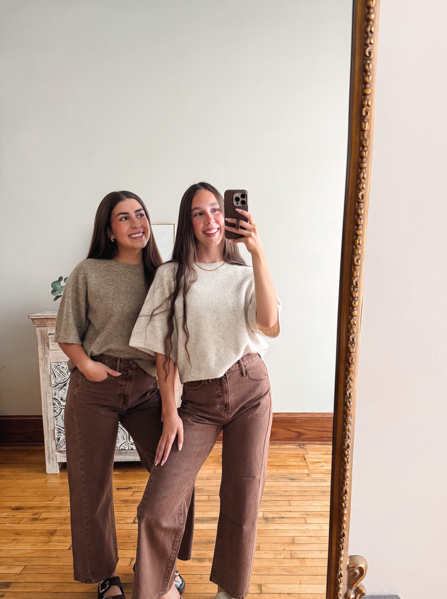 Two women taking a mirror selfie indoors, wearing matching brown wide-leg pants and neutral-colored short-sleeve tops. One wears a textured olive-green top and the other wears a light beige top.