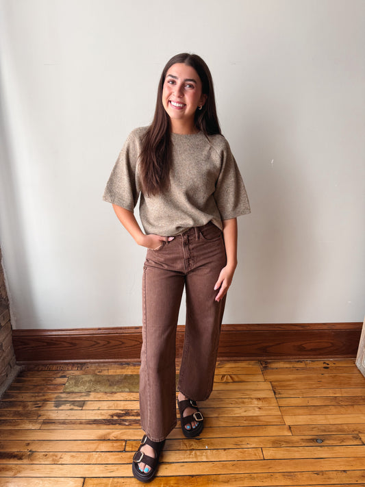 A woman modeling a short-sleeve taupe knit top and brown wide-leg jeans with black sandals, standing on a wooden floor against a plain off-white wall.