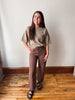 A woman modeling a short-sleeve taupe knit top and brown wide-leg jeans with black sandals, standing on a wooden floor against a plain off-white wall.