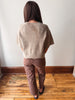 A person standing with their back to the camera, wearing a loose, short-sleeve knit sweater in a heathered taupe-beige color and brown pants, standing on a wooden floor against a plain wall.