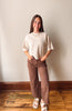 A woman stands indoors wearing a cream-colored short-sleeve knit top, brown wide-leg corduroy pants, and white slide sandals against a plain beige wall and wooden floor.