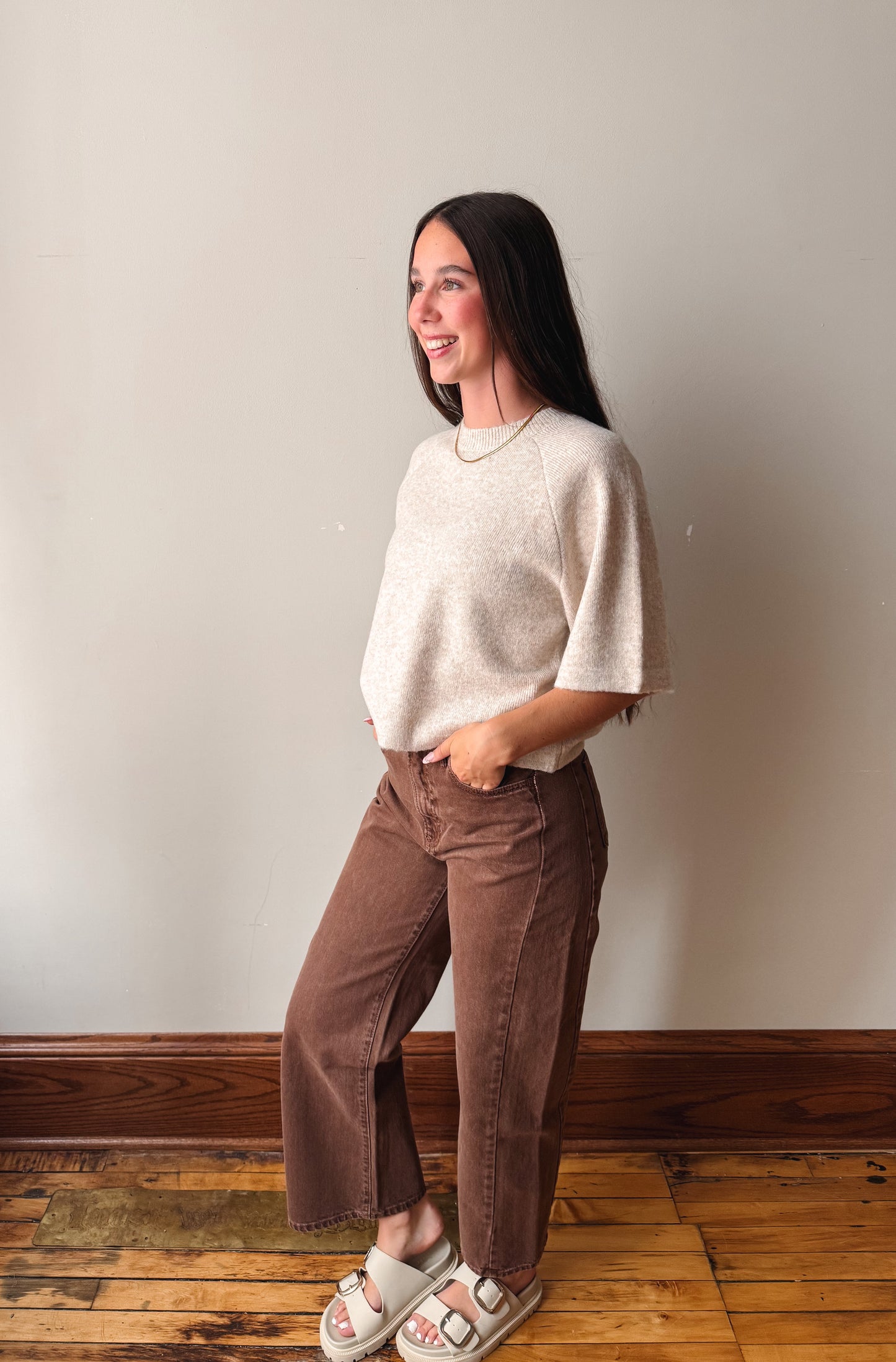 A woman modeling a cream-colored knit sweater and brown wide-leg pants, standing in a room with a light gray wall and wooden floor.