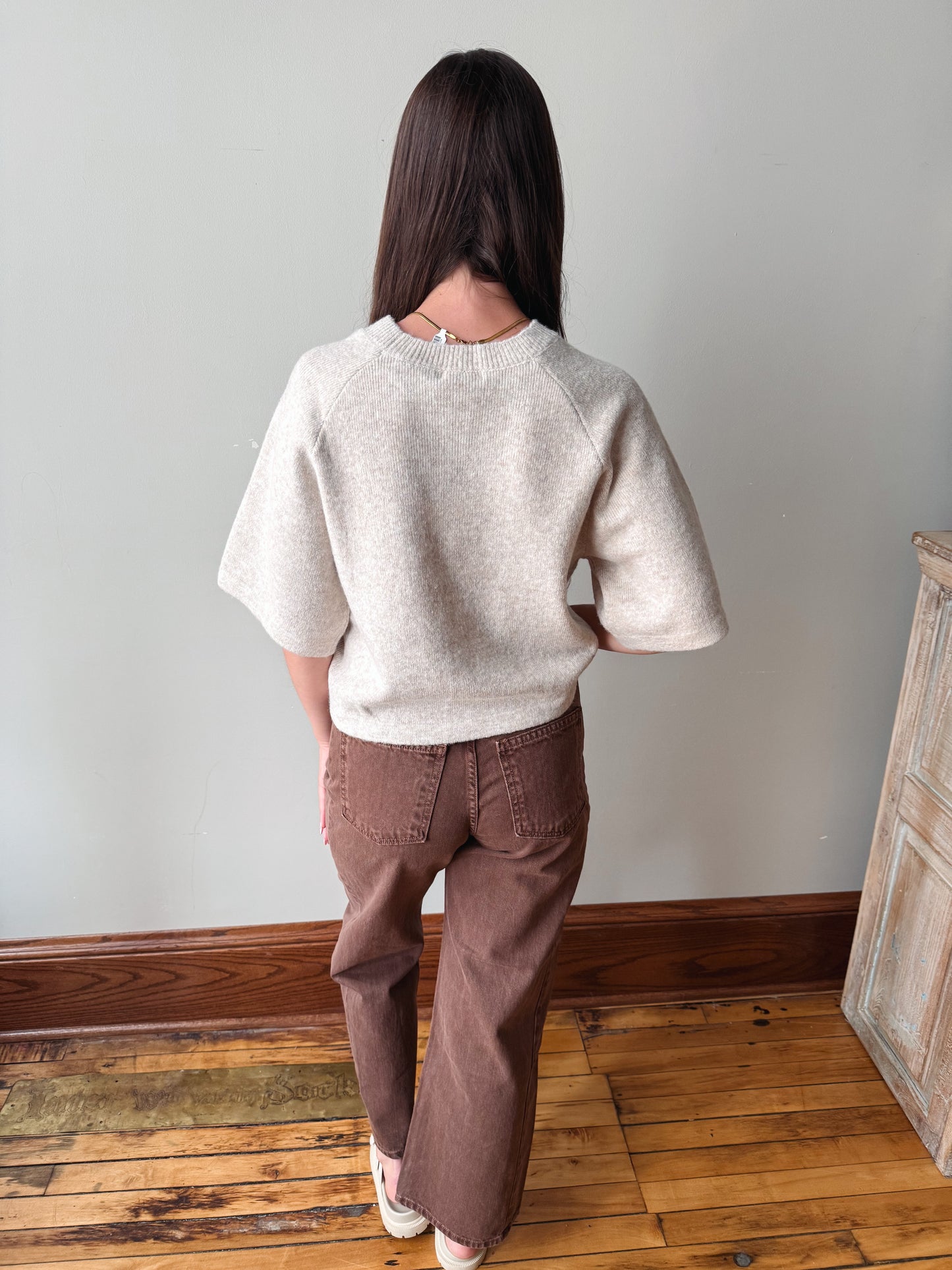 A woman viewed from behind wearing a light beige short-sleeve knit sweater and brown wide-leg pants, standing on a wooden floor against a plain light gray wall.