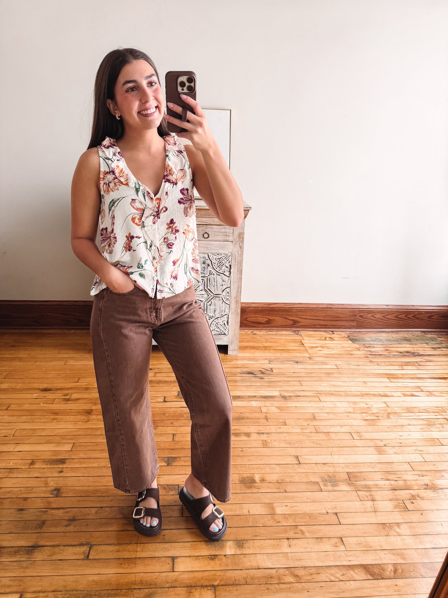A woman takes a mirror selfie in a room with hardwood floors and a white wall, wearing a sleeveless white top with a colorful floral print and brown cropped pants.