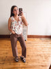 A woman takes a mirror selfie in a room with hardwood floors and a white wall, wearing a sleeveless white top with a colorful floral print and brown cropped pants.