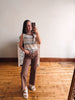 A woman takes a mirror selfie in a room with hardwood floors and a white wall, wearing a sleeveless striped tank top in beige, brown, and white, paired with brown wide-leg pants and white slide sandals.