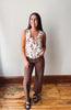 A woman modeling a sleeveless wrap-style blouse with a multicolor floral print on a cream background, paired with brown wide-leg pants and black slide sandals, standing in a room with a light gray wall and wooden floor.