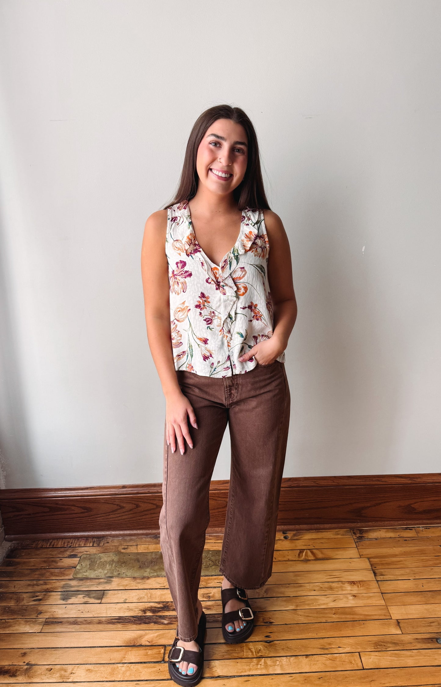 A woman modeling a sleeveless white top with a colorful floral print and brown wide-leg pants, standing on a wooden floor against a plain wall.