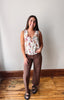 A woman modeling a sleeveless white top with a colorful floral print and brown wide-leg pants, standing on a wooden floor against a plain wall.