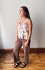A woman modeling a sleeveless white top with a colorful butterfly and floral print, paired with brown wide-leg pants and black sandals, standing on a wooden floor against a plain wall.