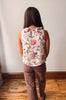 A woman viewed from behind wearing a sleeveless white top with a colorful floral print and brown pants, standing on a wooden floor against a plain wall.