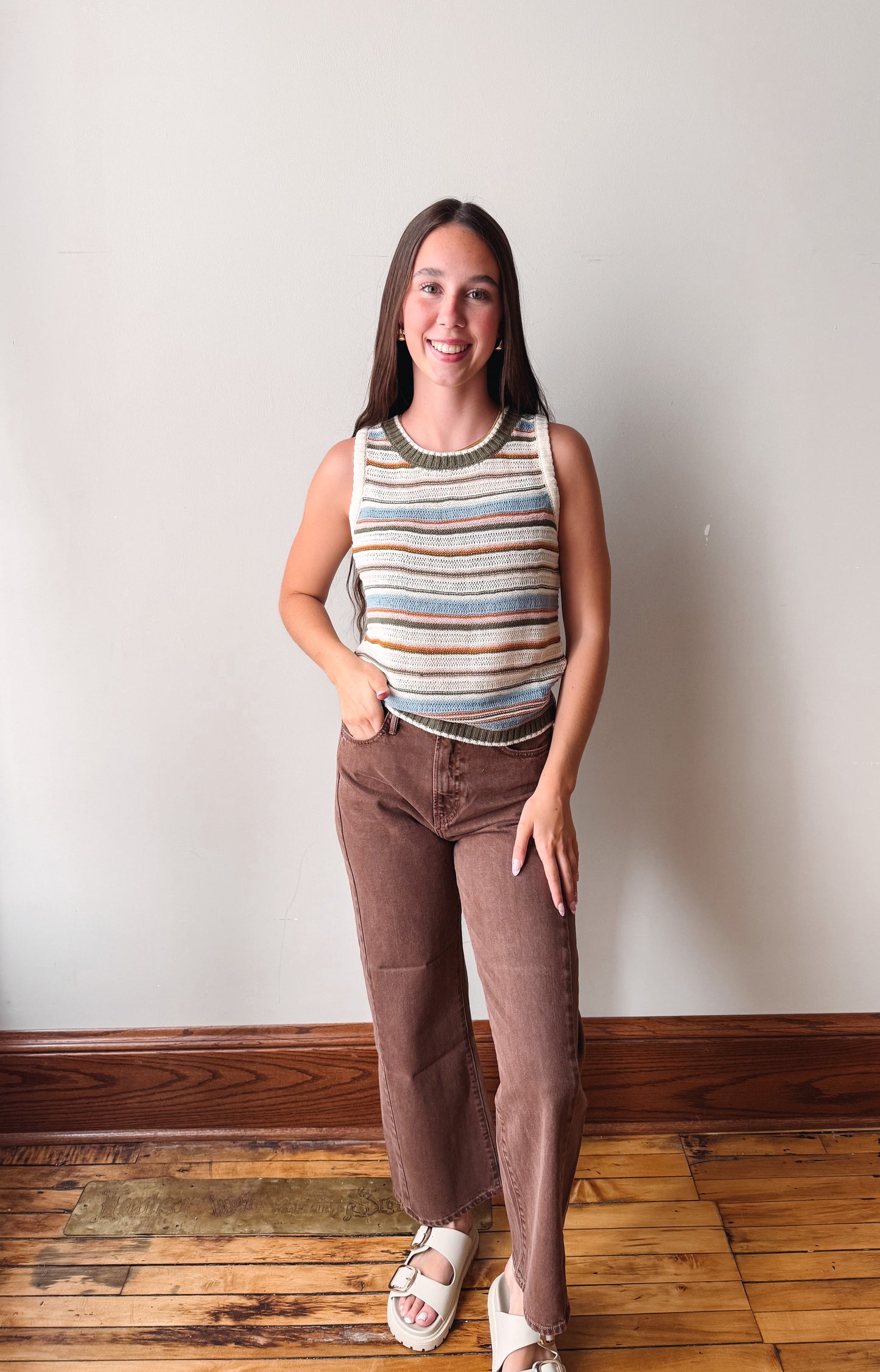 A woman stands in a room with a light gray wall and wooden floor, wearing a sleeveless knit top with horizontal stripes in beige, brown, and blue, paired with brown pants and white slide sandals.