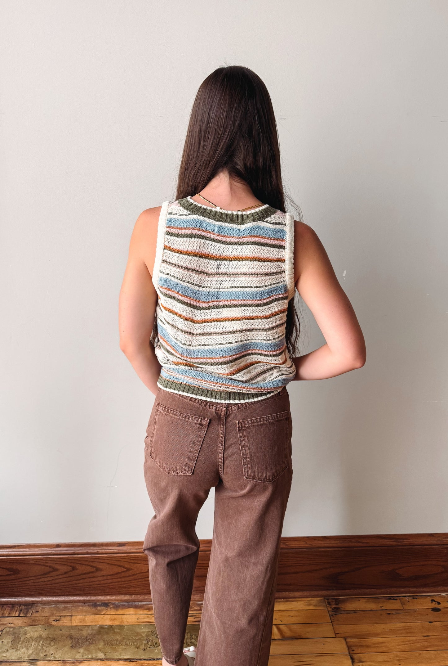 A woman viewed from behind wearing a sleeveless knit top with horizontal stripes in blue, beige, brown, and white, paired with brown pants, standing against a plain light gray wall.