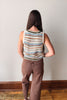 A woman viewed from behind wearing a sleeveless knit top with horizontal stripes in blue, beige, brown, and white, paired with brown pants, standing against a plain light gray wall.