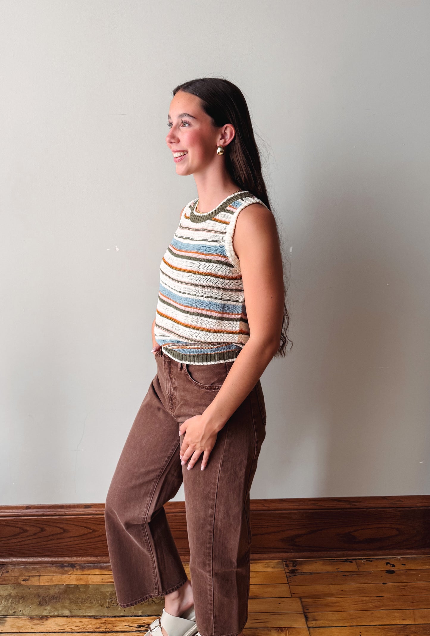 A woman modeling a sleeveless knit top with horizontal stripes in cream, blue, brown, and orange, paired with brown corduroy pants, standing in a room with a light gray wall and wooden floor.