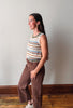 A woman modeling a sleeveless knit top with horizontal stripes in cream, blue, brown, and orange, paired with brown corduroy pants, standing in a room with a light gray wall and wooden floor.