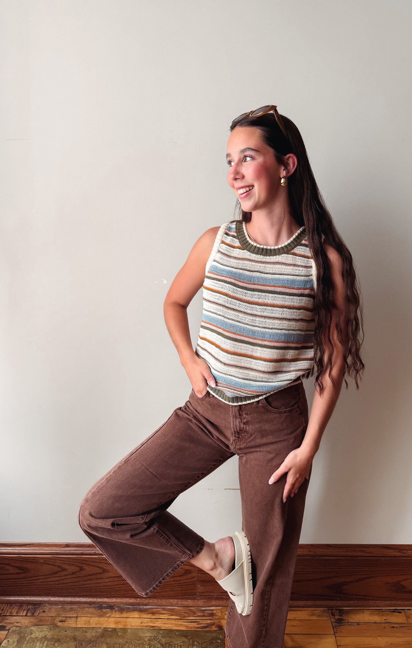 A woman posing in a striped sleeveless knit top with brown wide-leg jeans and white sneakers, standing against a plain off-white wall with wooden baseboards.
