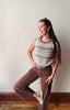 A woman posing in a striped sleeveless knit top with brown wide-leg jeans and white sneakers, standing against a plain off-white wall with wooden baseboards.