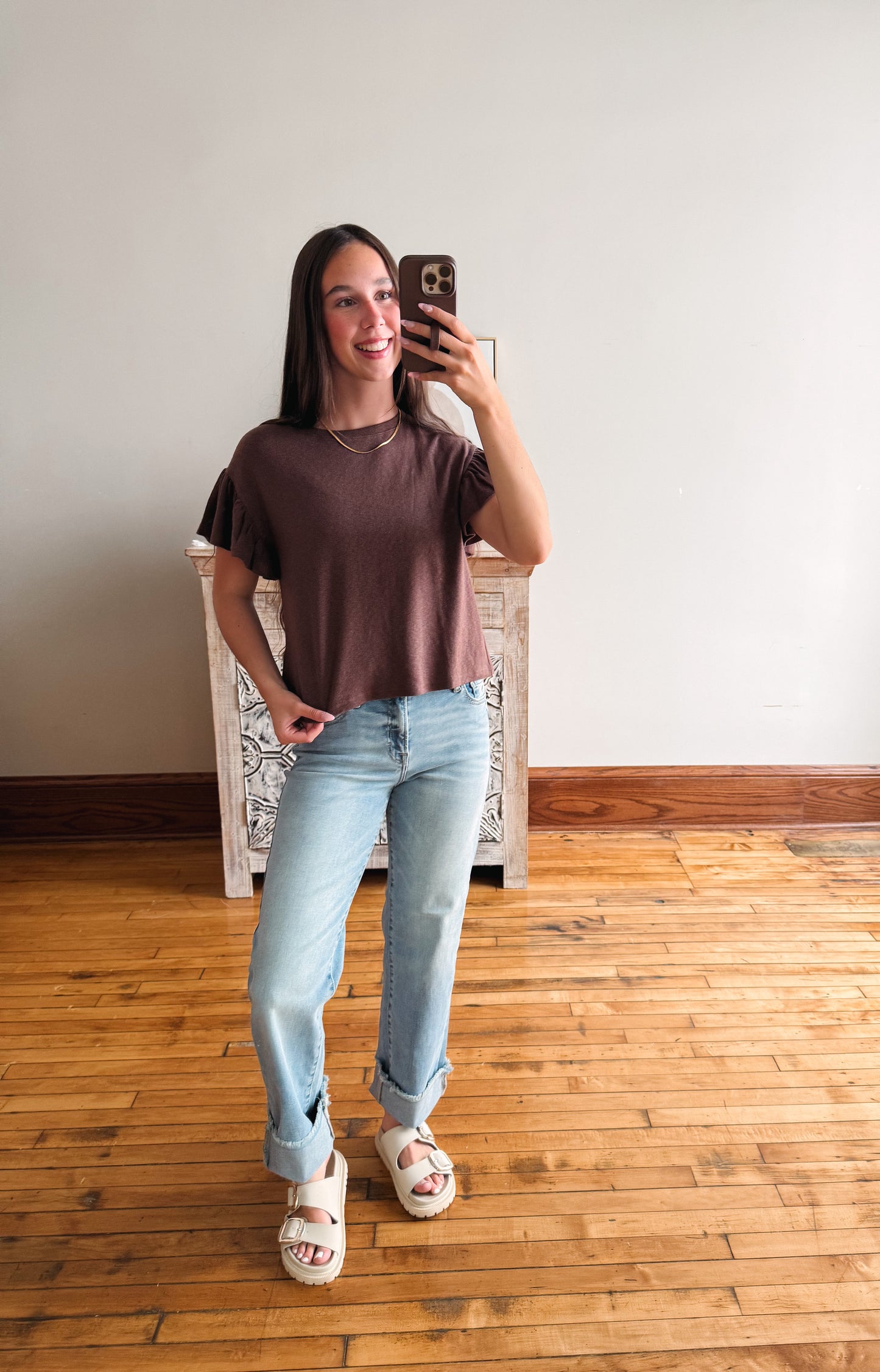 A woman takes a mirror selfie wearing a brown short-sleeve t-shirt, light wash blue jeans, and white slide sandals in a room with a wooden floor and white wall.