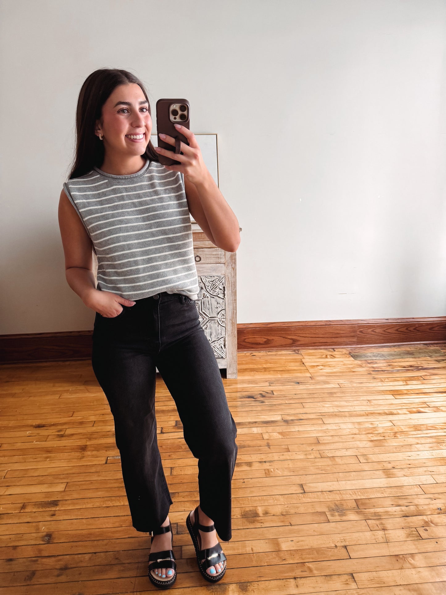 A woman takes a mirror selfie in a room with hardwood floors and a white wall, wearing a gray and white striped sleeveless top, black cropped pants, and black sandals.