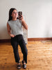 A woman takes a mirror selfie in a room with hardwood floors and a white wall, wearing a gray and white striped sleeveless top, black cropped pants, and black sandals.