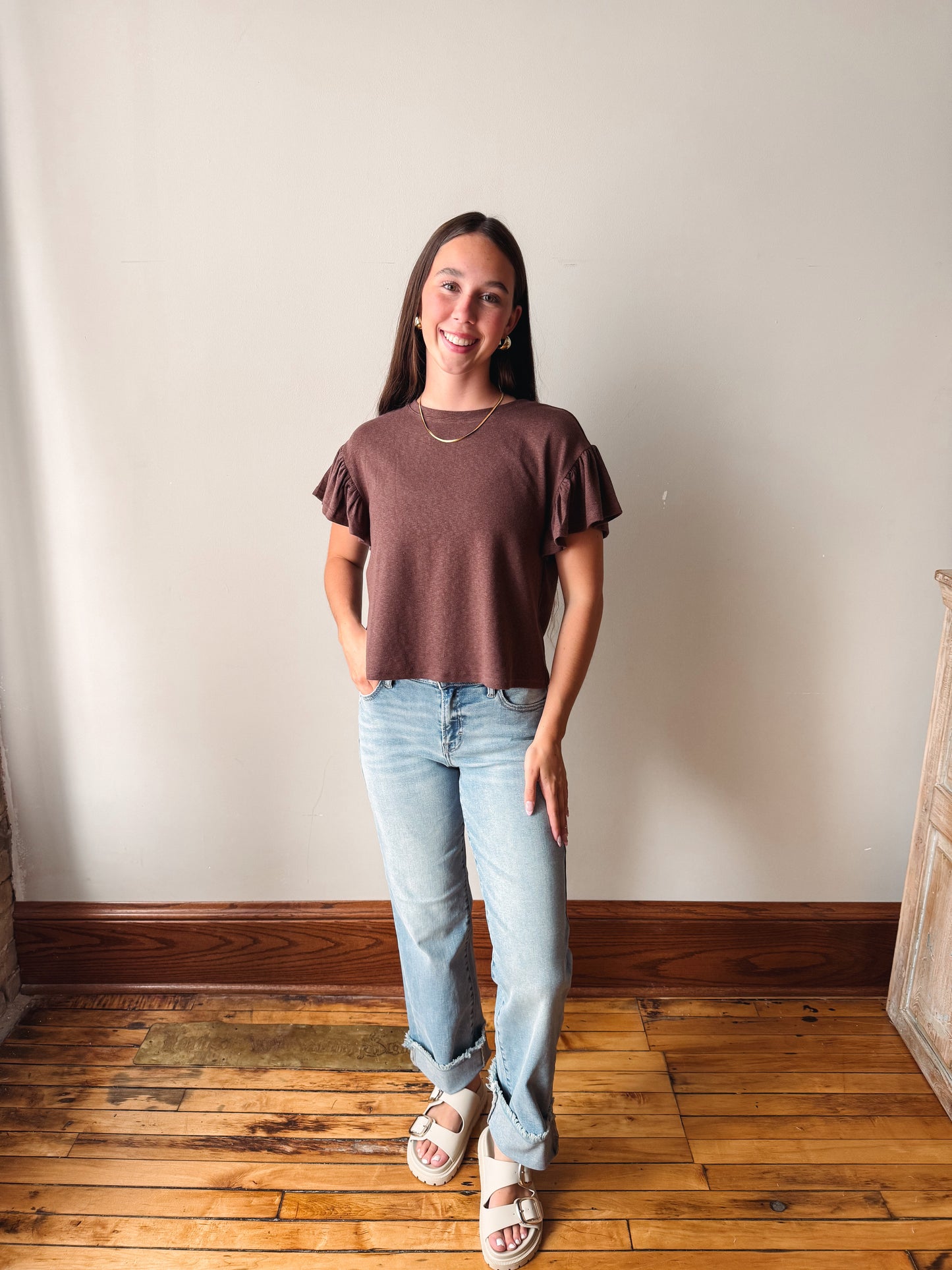 A woman stands in a room with a beige wall and wooden floor, wearing a short-sleeve ruffled-sleeve top in a muted brownish-purple color, light-wash blue jeans, and white slide sandals.