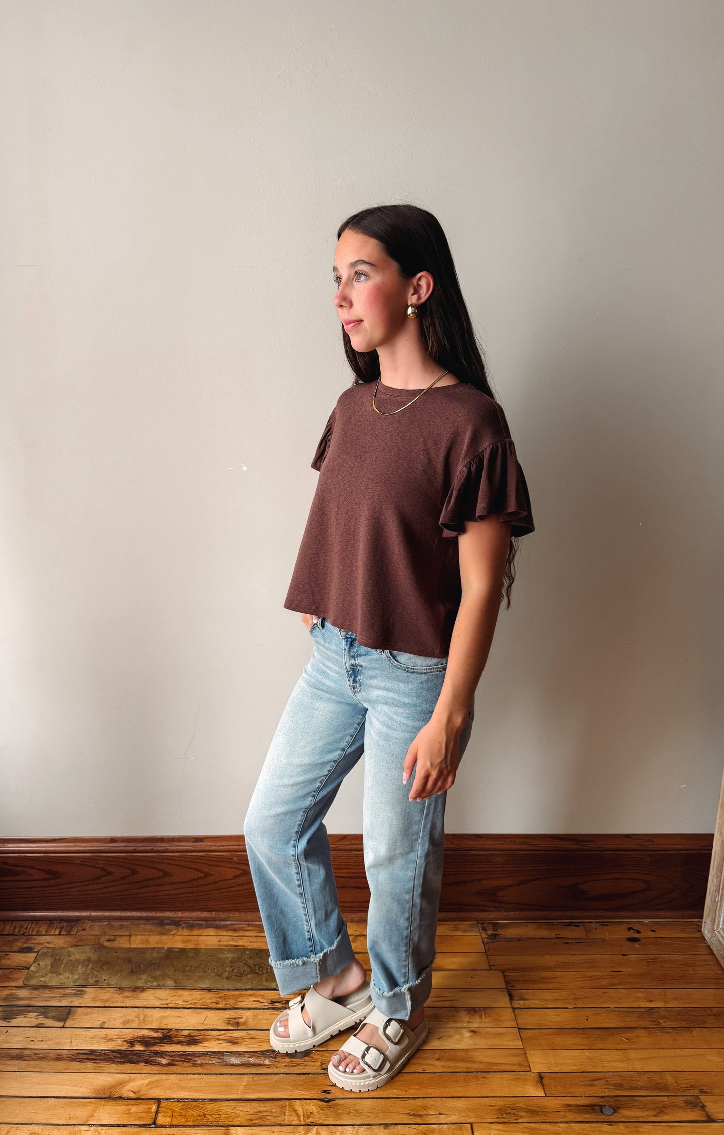 A woman modeling a brown short-sleeve crop top with ruffled sleeves, paired with light blue jeans and white slide sandals, standing in a room with a beige wall and wooden floor.