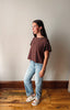 A woman modeling a brown short-sleeve crop top with ruffled sleeves, paired with light blue jeans and white slide sandals, standing in a room with a beige wall and wooden floor.