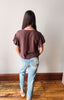 A woman viewed from behind wearing a dark brown ruffle-sleeve t-shirt and light blue jeans, standing on a wooden floor against a plain wall.